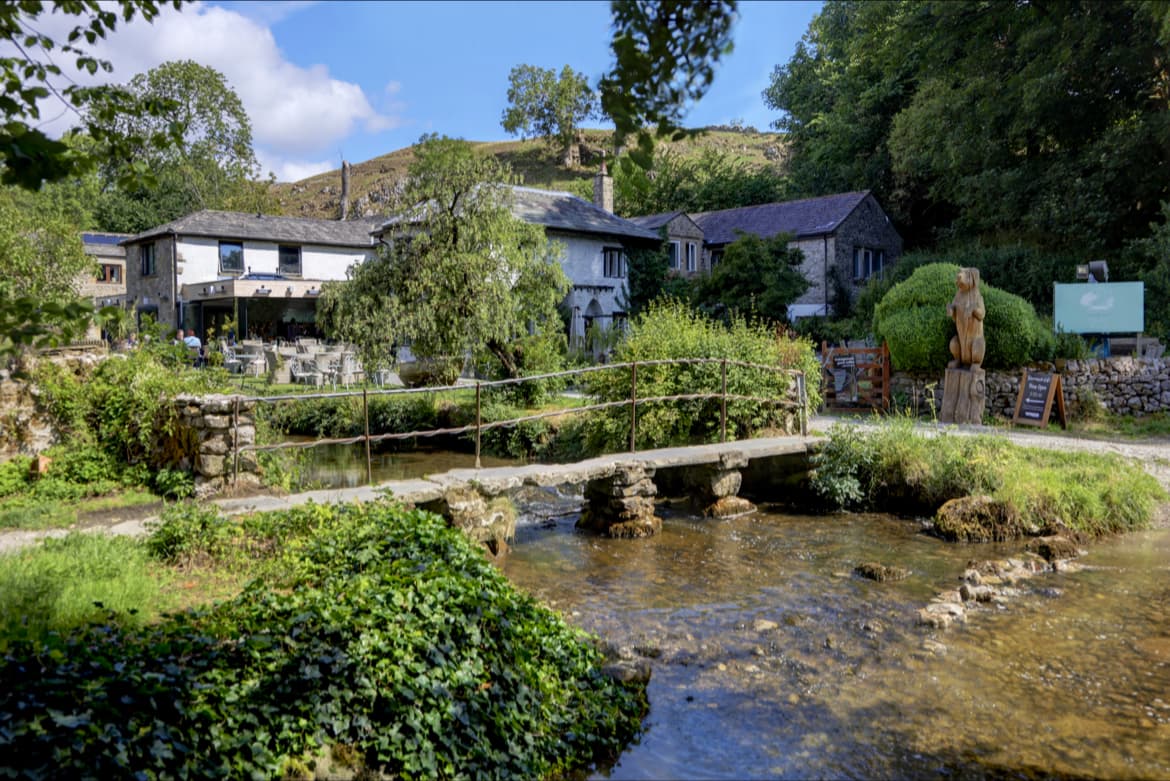 Charming stone cafe with outdoor seating, a small bridge over a stream, and lush greenery in Kirkby Malham, Yorkshire.