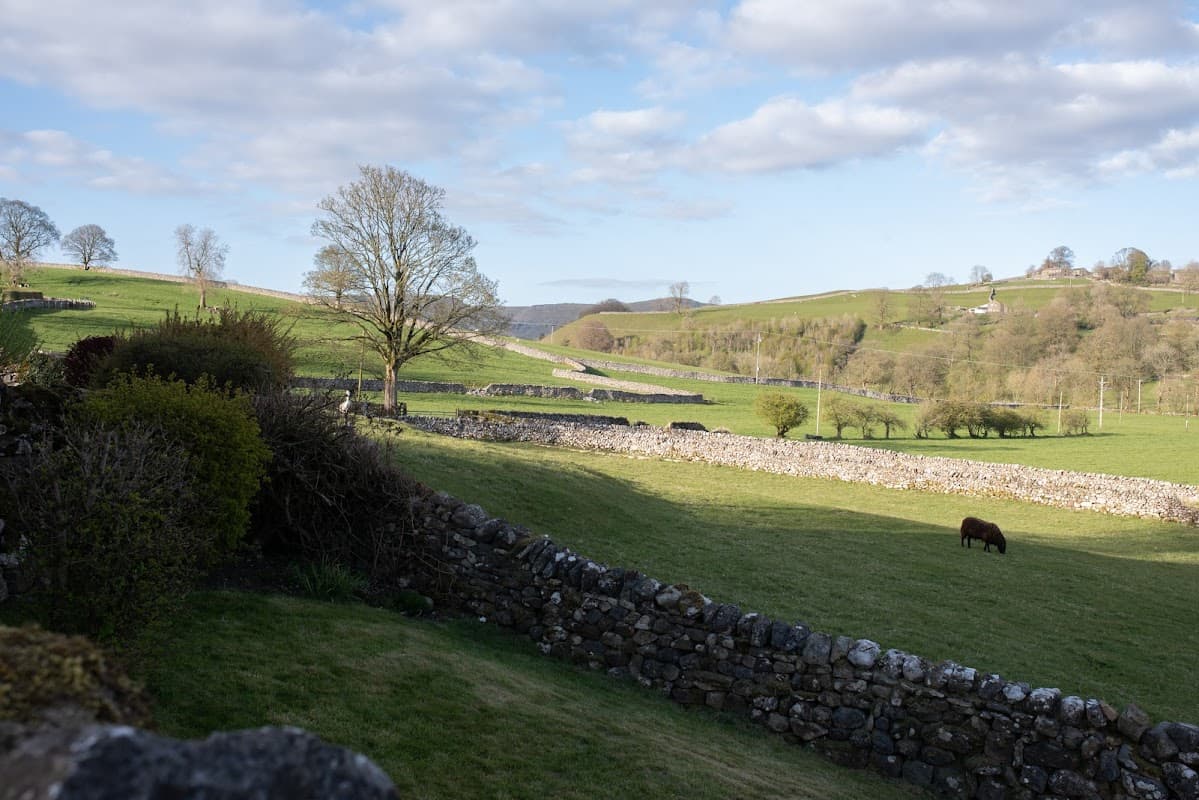 Lush green fields and dry stone walls under a blue sky with scattered clouds, featuring a grazing horse in the foreground.