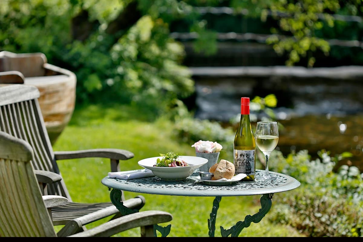 A table set with a salad, bread, a bottle of wine, and a glass, surrounded by greenery near a stream.