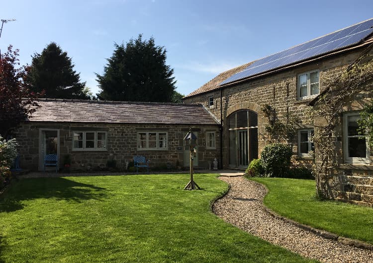 Charming stone cottage with solar panels, surrounded by lush greenery and a gravel path in Kirkby Malzeard, Yorkshire.