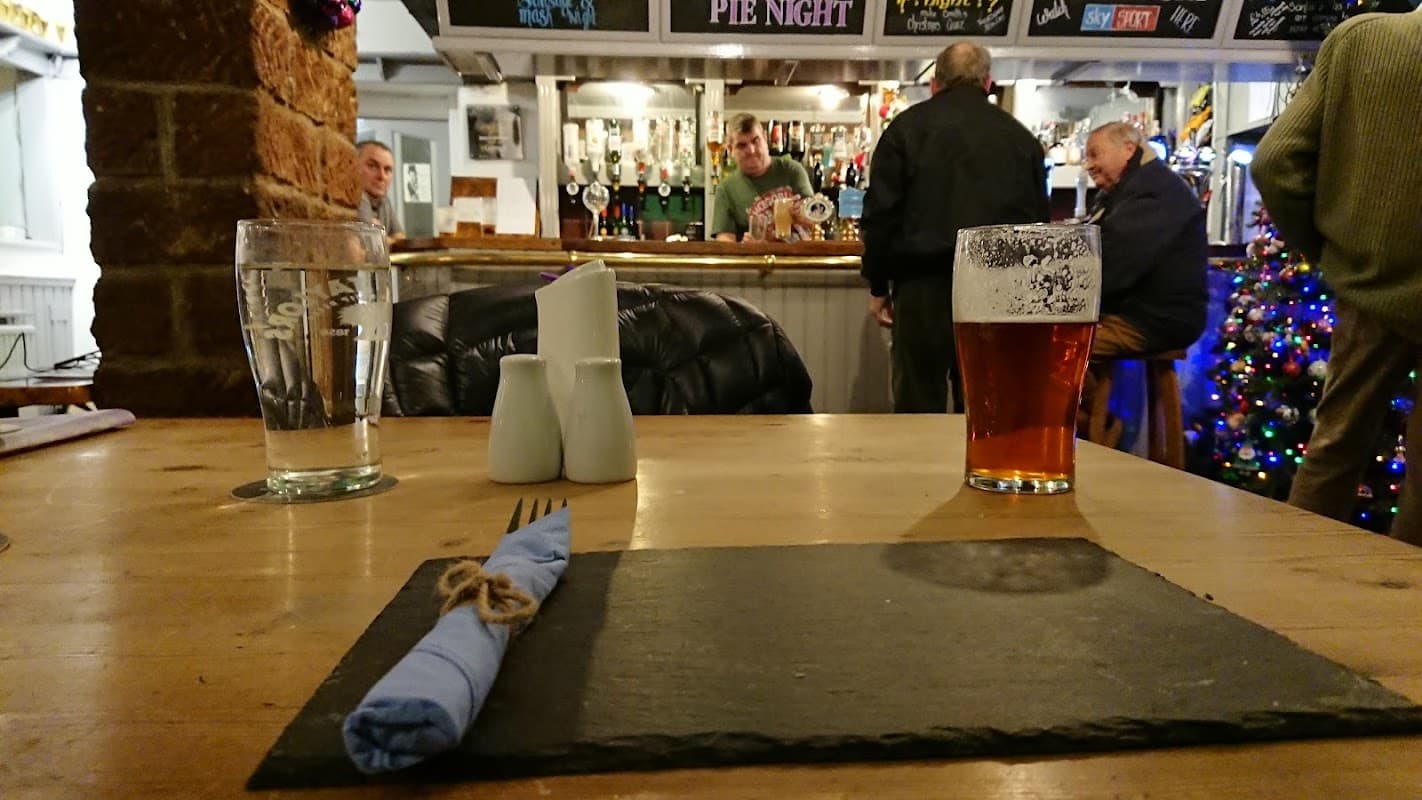 A table set with a slate placemat, cutlery, a pint of beer, and a view of a bustling bar with patrons.