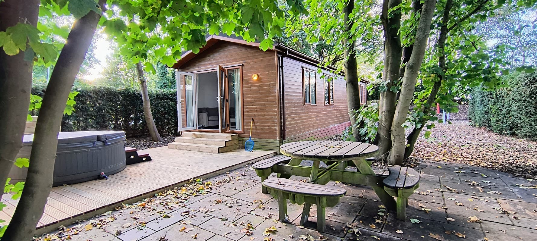 Wooden cabin surrounded by trees, with a hot tub and picnic table on a stone patio.