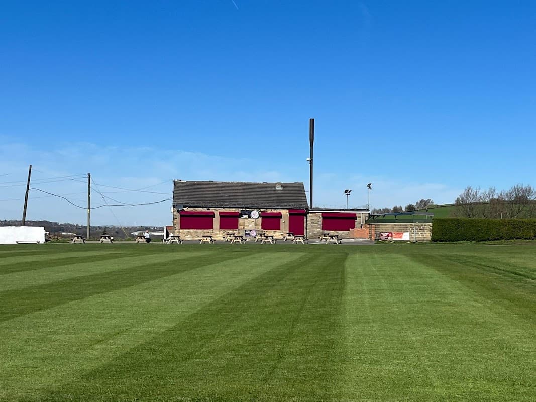 Kirkheaton Cricket and Bowling Club with a well-maintained green field and a brick building under a clear blue sky.