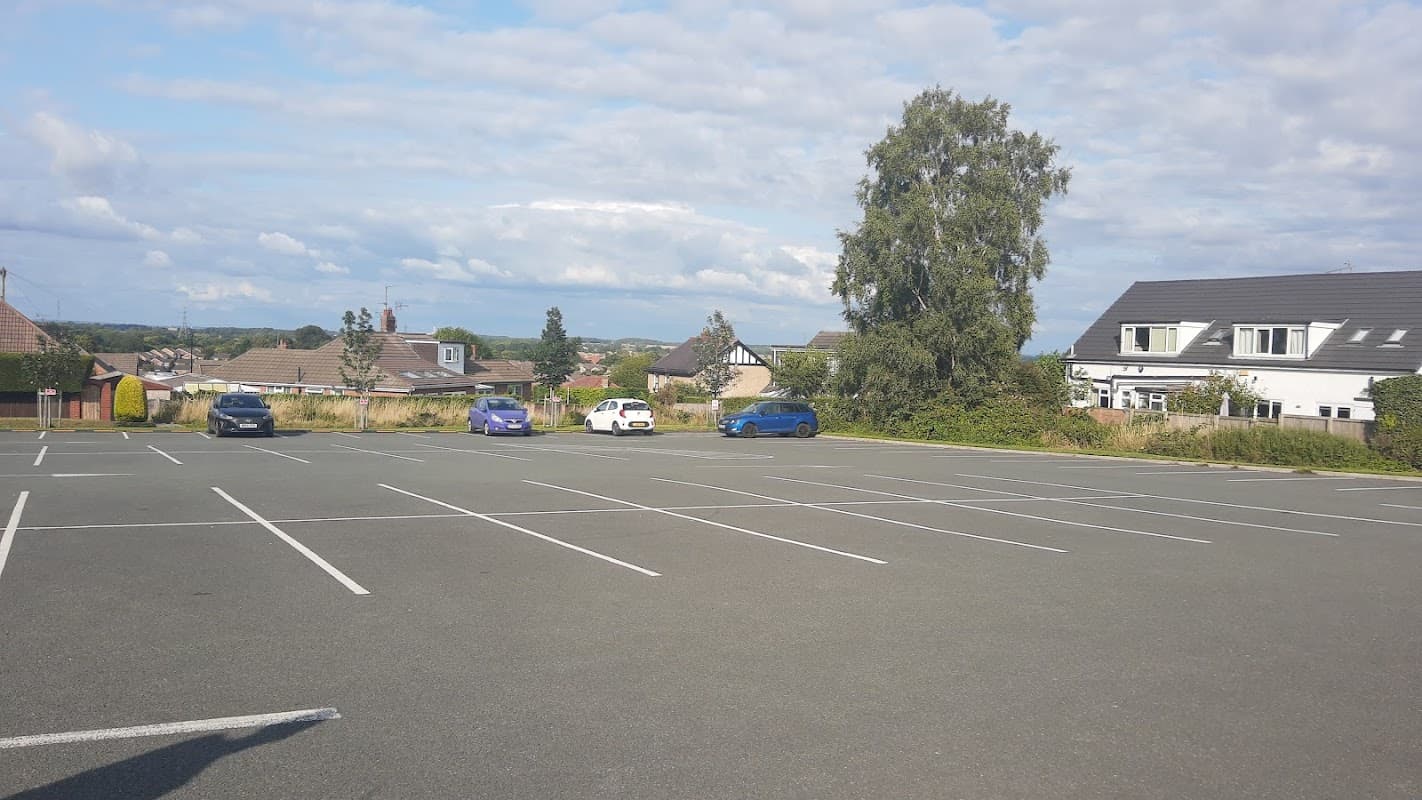 Empty car park with a few parked cars, surrounded by greenery and residential buildings under a cloudy sky.