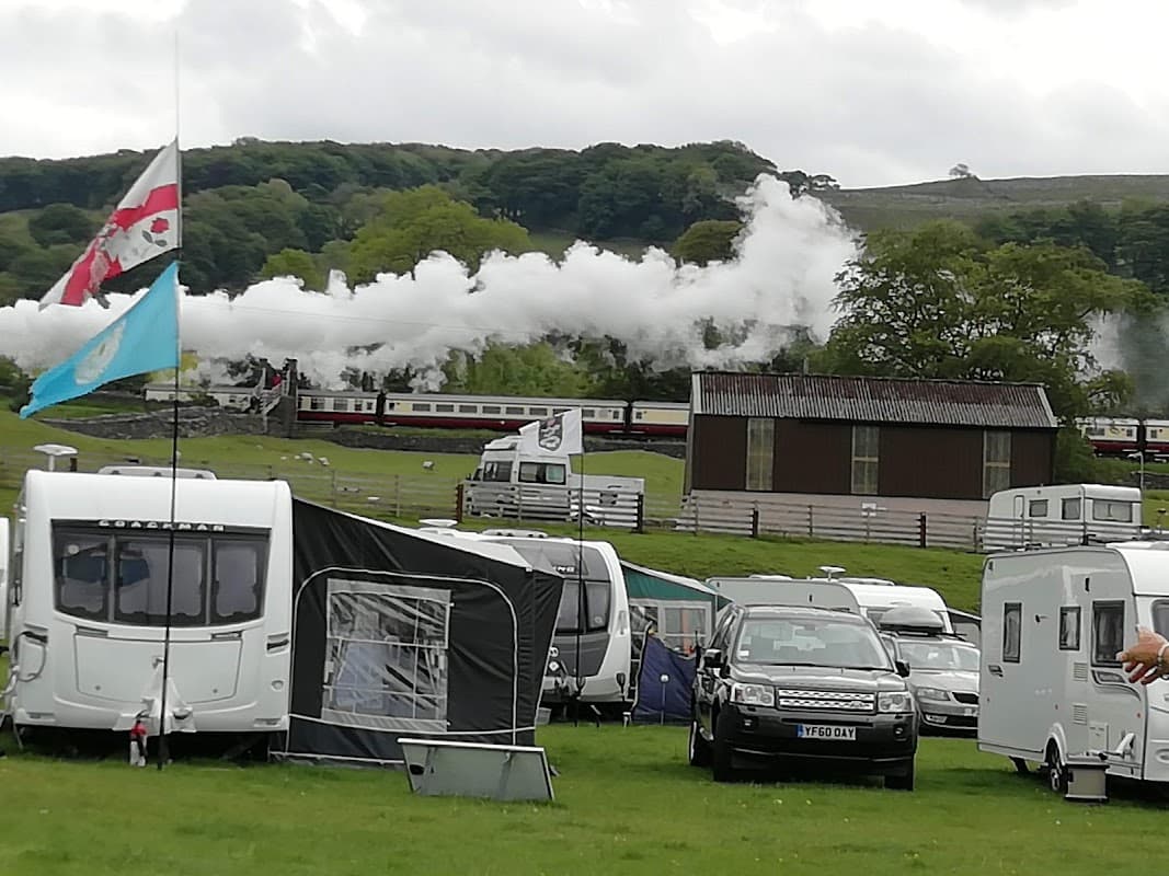 Steam train chugs through green landscape with caravans and tents in the foreground, set in Langcliffe, Yorkshire.