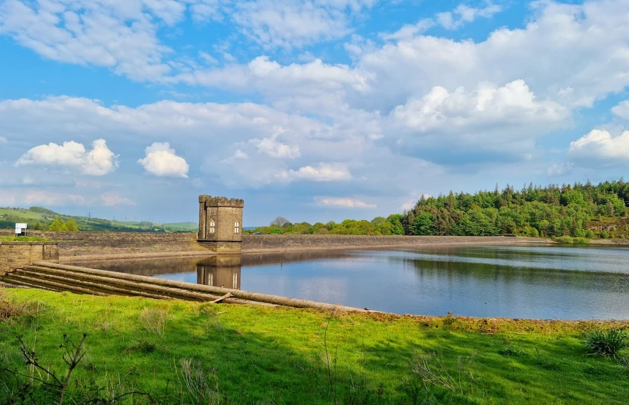 A serene landscape featuring a reservoir with a stone tower, surrounded by lush greenery and blue sky with clouds.