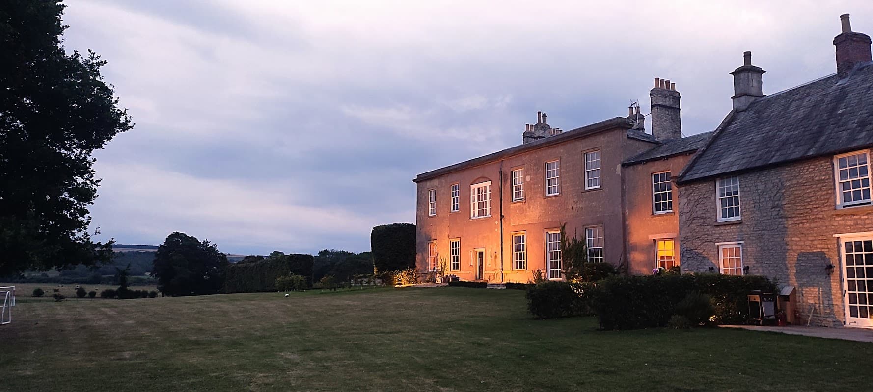 Langton Hall features a stone exterior with warm lighting, surrounded by a manicured lawn and trees under a cloudy sky.