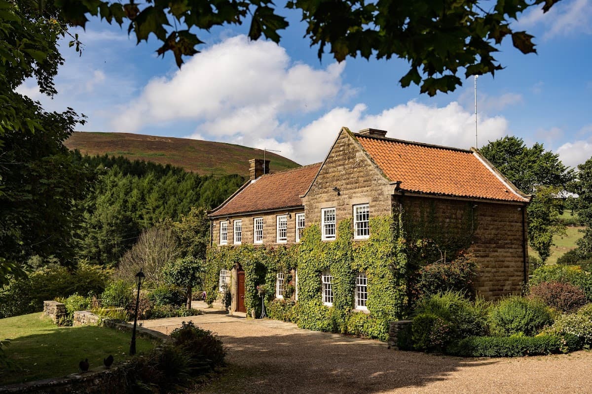 Stone country house covered in greenery, surrounded by gardens and hills under a partly cloudy sky.