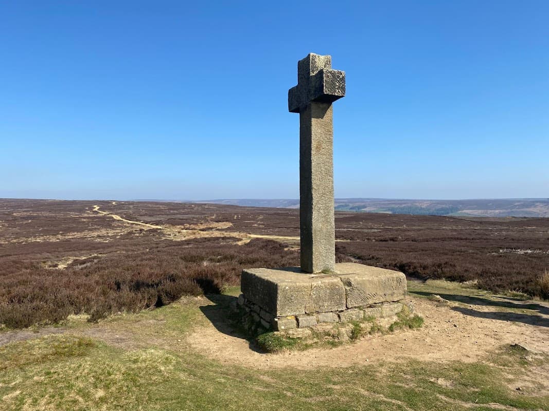 Stone cross on a grassy hilltop with heather-covered landscape and clear blue sky in Lastingham, Yorkshire.