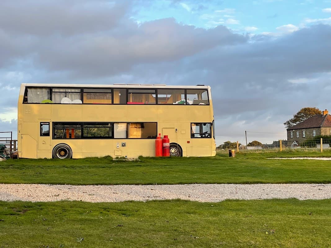 A converted double-decker bus sits on green grass, surrounded by a scenic landscape in Laverton, North Yorkshire.