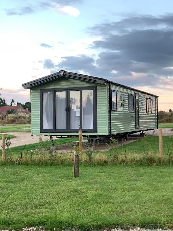 Green holiday home on a grassy area at Broadbeck Holiday Park, with large windows and a cloudy sky in the background.