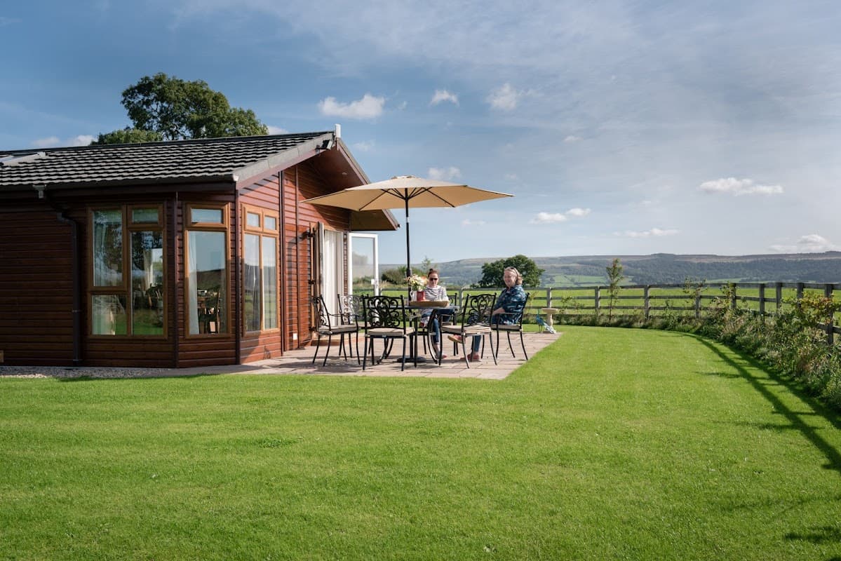 A wooden lodge with a patio, umbrella, and two people enjoying the view of green fields and hills in the background.