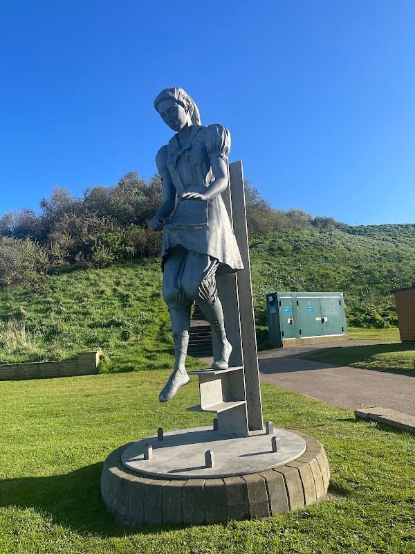 A large statue of a girl in a dress, standing on a pedestal, surrounded by green grass and a clear blue sky.