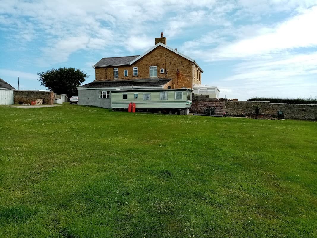 A green caravan on a grassy field beside a large stone house under a blue sky with scattered clouds.