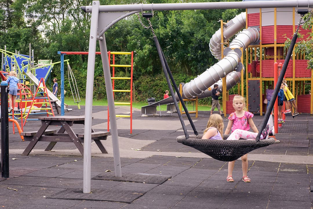 Children playing on a swing and near colorful playground equipment with a slide and climbing structures in a park setting.