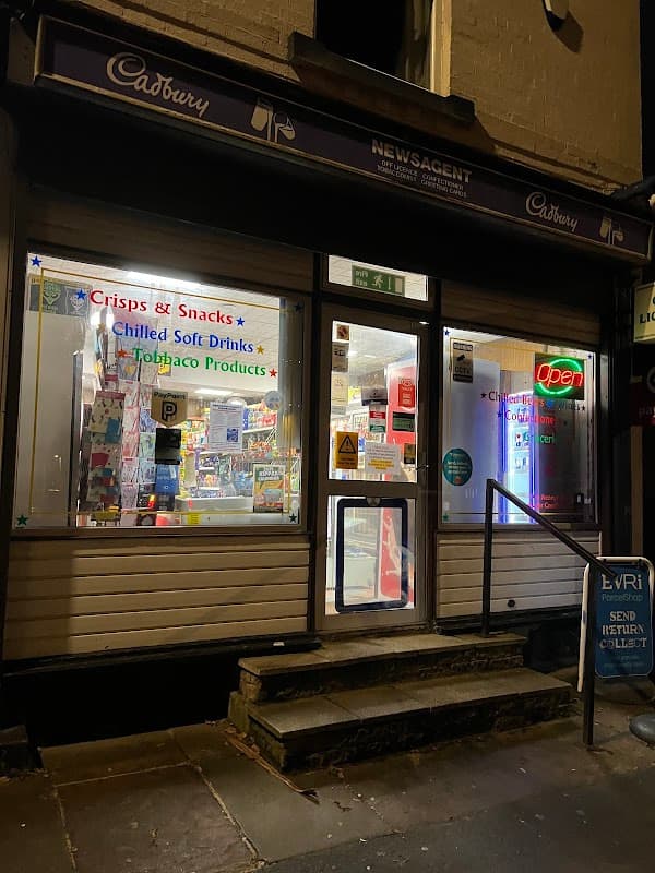 Newsagent storefront with illuminated signs, displaying snacks, drinks, and tobacco products, in a corner shop setting.