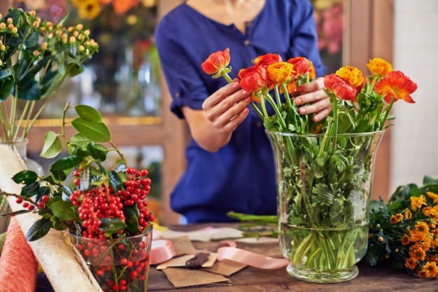A florist arranging vibrant orange and yellow flowers in a glass vase, surrounded by greenery and decorative materials.