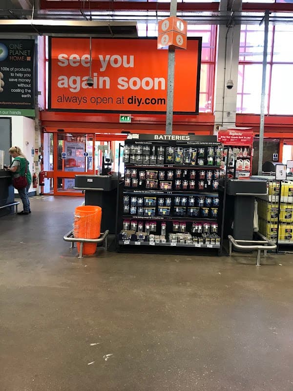 B&Q store interior with battery display, checkout area, and a large sign reading "see you again soon."