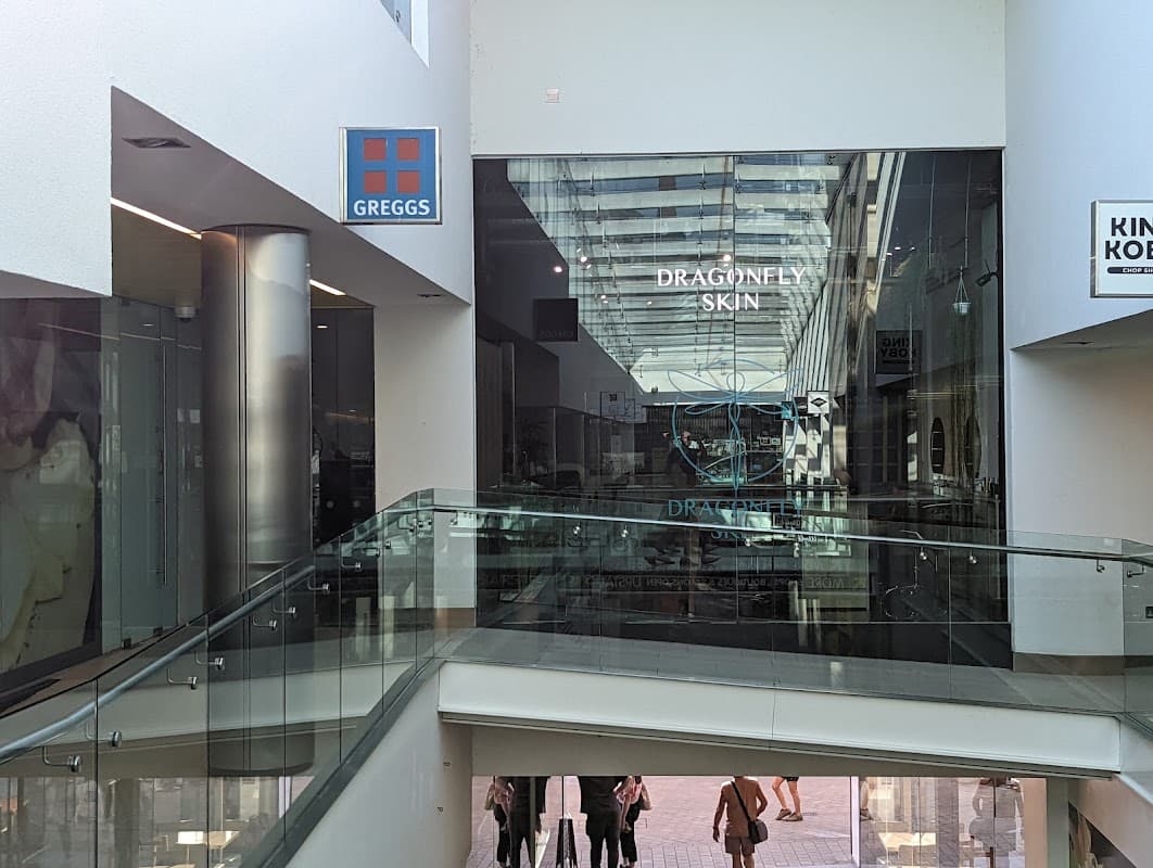 Central Arcade interior featuring glass railings, shops, and signage for Greggs and Dragonfly Skin.