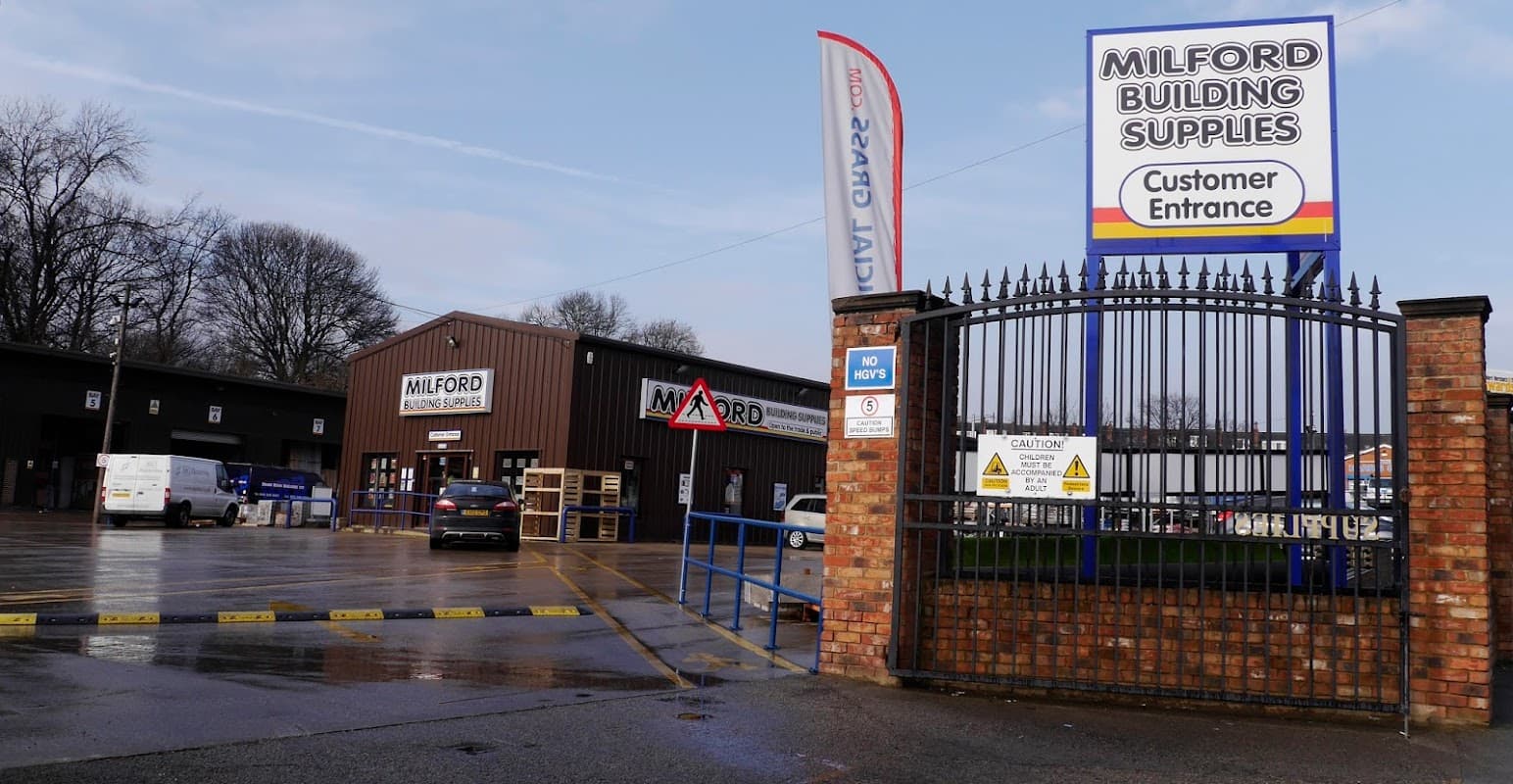 Milford Building Supplies entrance with a sign, vehicles in the lot, and trees in the background. Wet pavement visible.