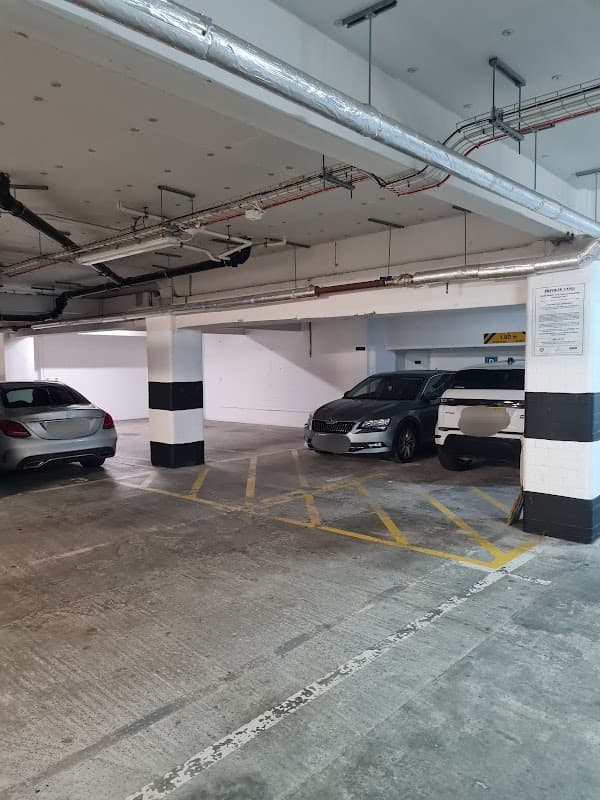 Empty parking area with two parked cars, concrete flooring, and overhead pipes in a well-lit garage.