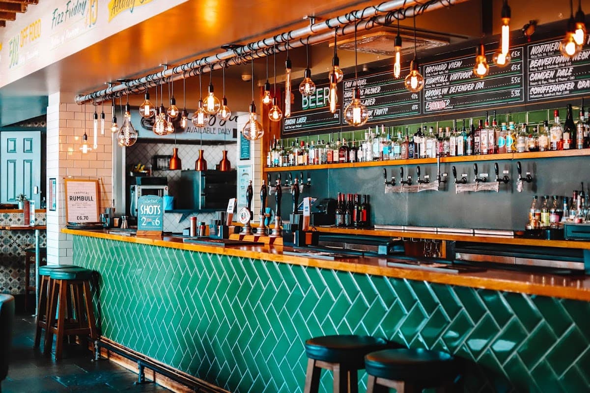 A vibrant bar with green tiled counter, hanging lights, and shelves stocked with various bottles and taps.