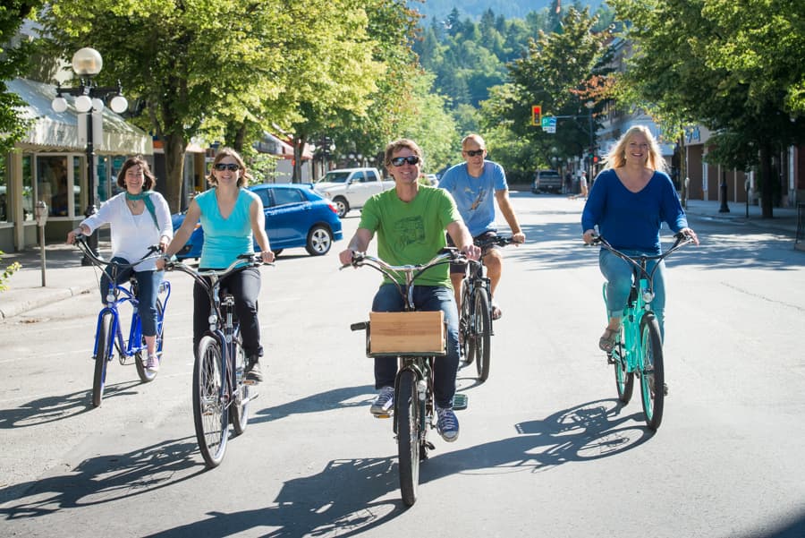 A group of five people riding bicycles on a sunny street, surrounded by trees and shops.