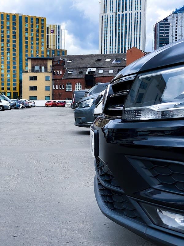 Cars parked in a spacious lot with modern buildings and a cloudy sky in the background.