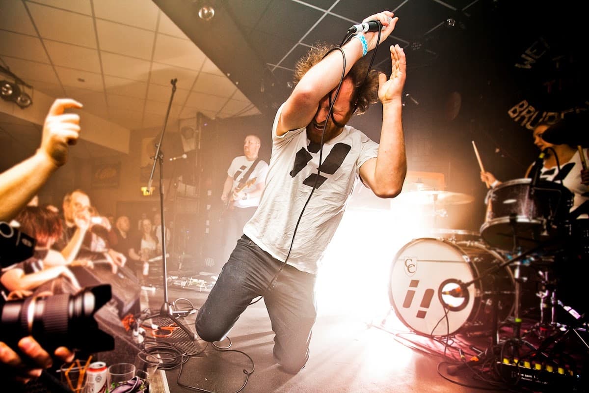 A performer kneels on stage, singing passionately, with a lively crowd and band in the background.