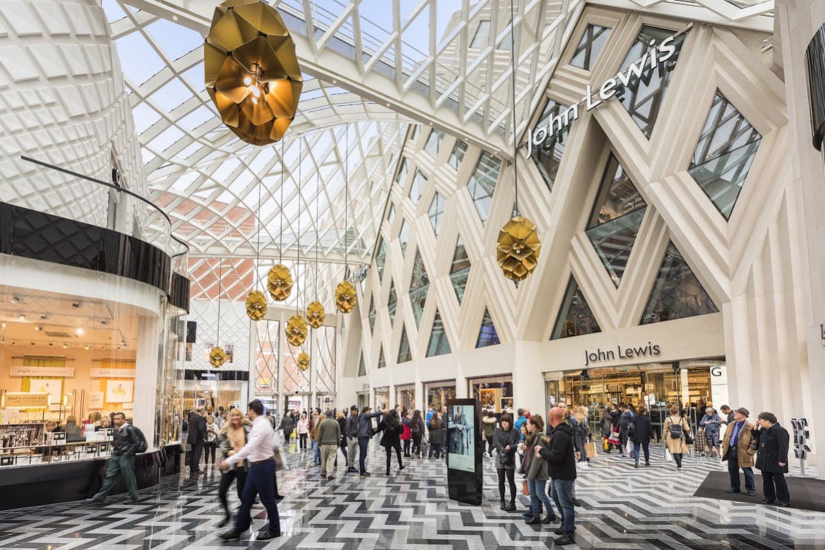 Modern shopping center with a glass roof, geometric patterns on the floor, and shoppers exploring various stores.
