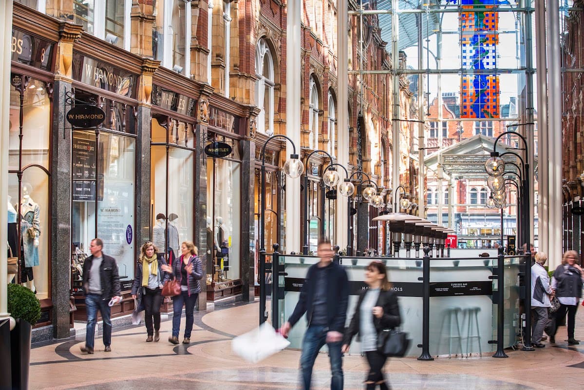 Victorian-style shopping arcade with glass roof, ornate architecture, and shoppers walking through the bustling space.
