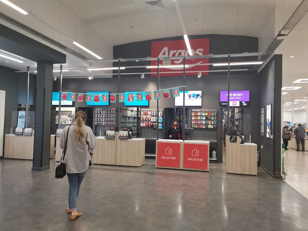 Interior of a retail space with an Argos collection counter, digital screens, and a customer browsing.