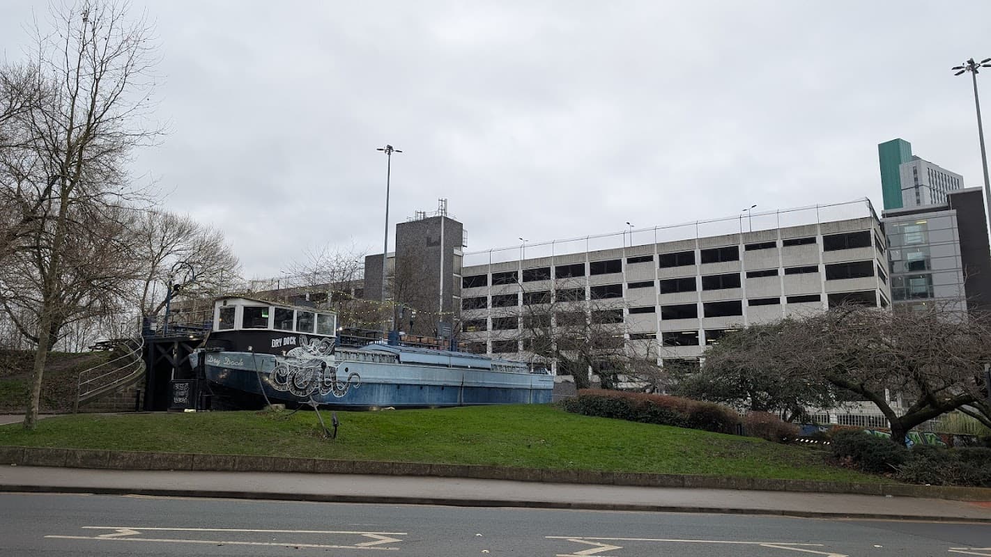 Multi-storey car park with a blue boat on grass in front, surrounded by trees and urban buildings in Leeds.