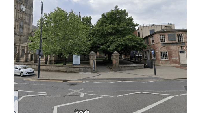 A street view of a park entrance with large trees, historic stone pillars, and nearby brick buildings in Leeds.