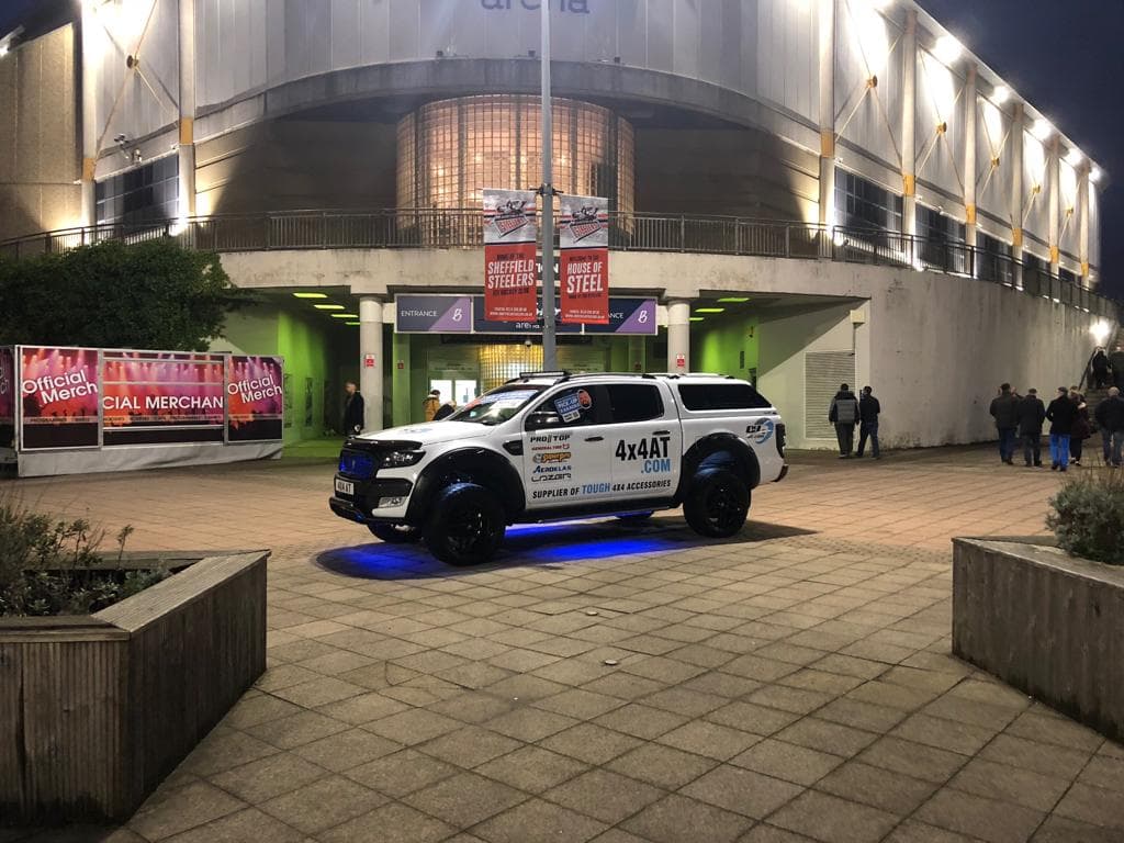 A modified 4x4 vehicle parked near an arena, illuminated with blue lights, showcasing branding for 4x4 Accessories and Tyres.