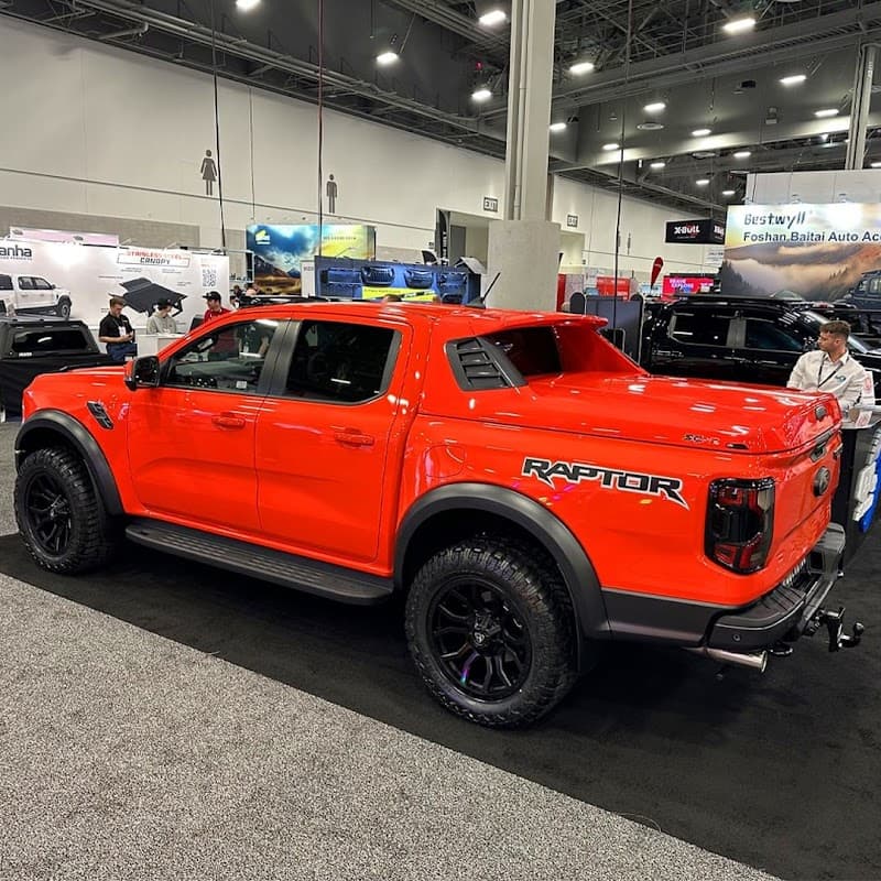 Bright orange Ford Raptor truck showcased at a car parts exhibition, featuring rugged tires and a sporty design.