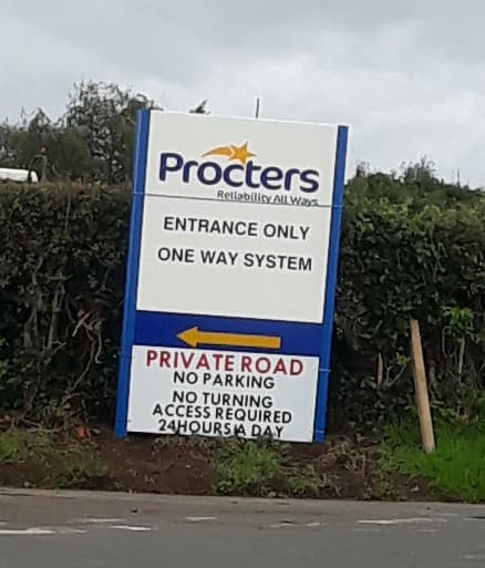 Procters Coaches sign with "Entrance Only," "One Way System," and "Private Road" instructions, surrounded by greenery.
