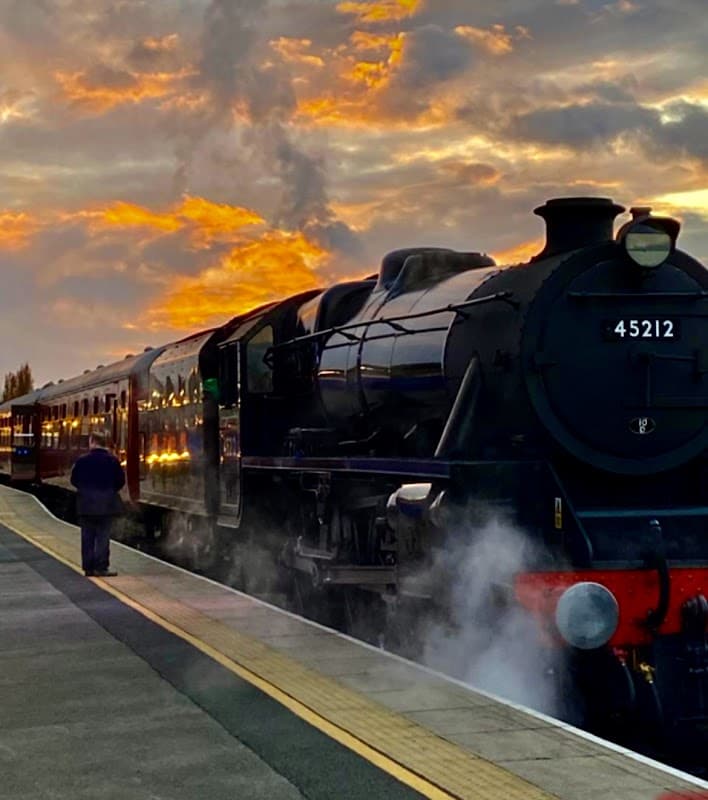 Steam train 45212 at Wensleydale Railway station, with a dramatic sunset and a person standing on the platform.