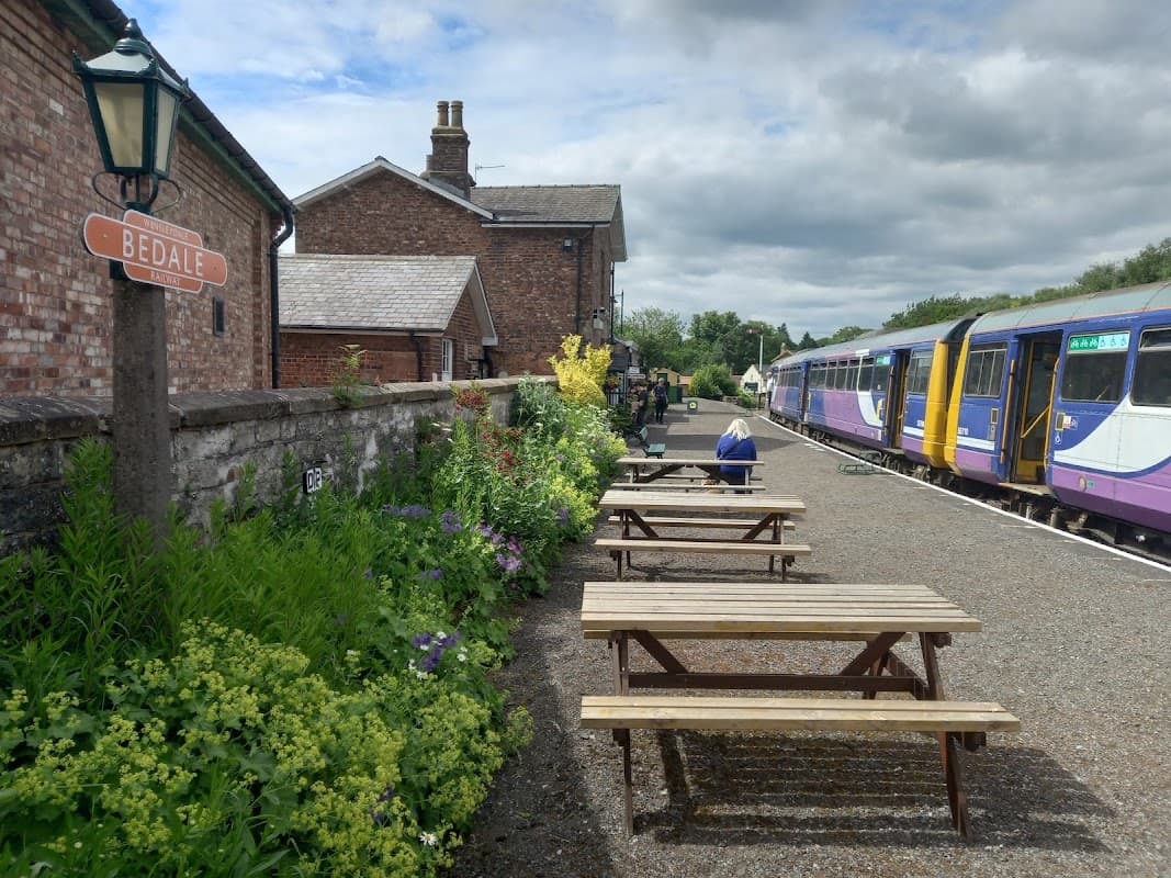 Bedale Station features a sign, flowerbeds, wooden benches, and trains at the platform under a cloudy sky.