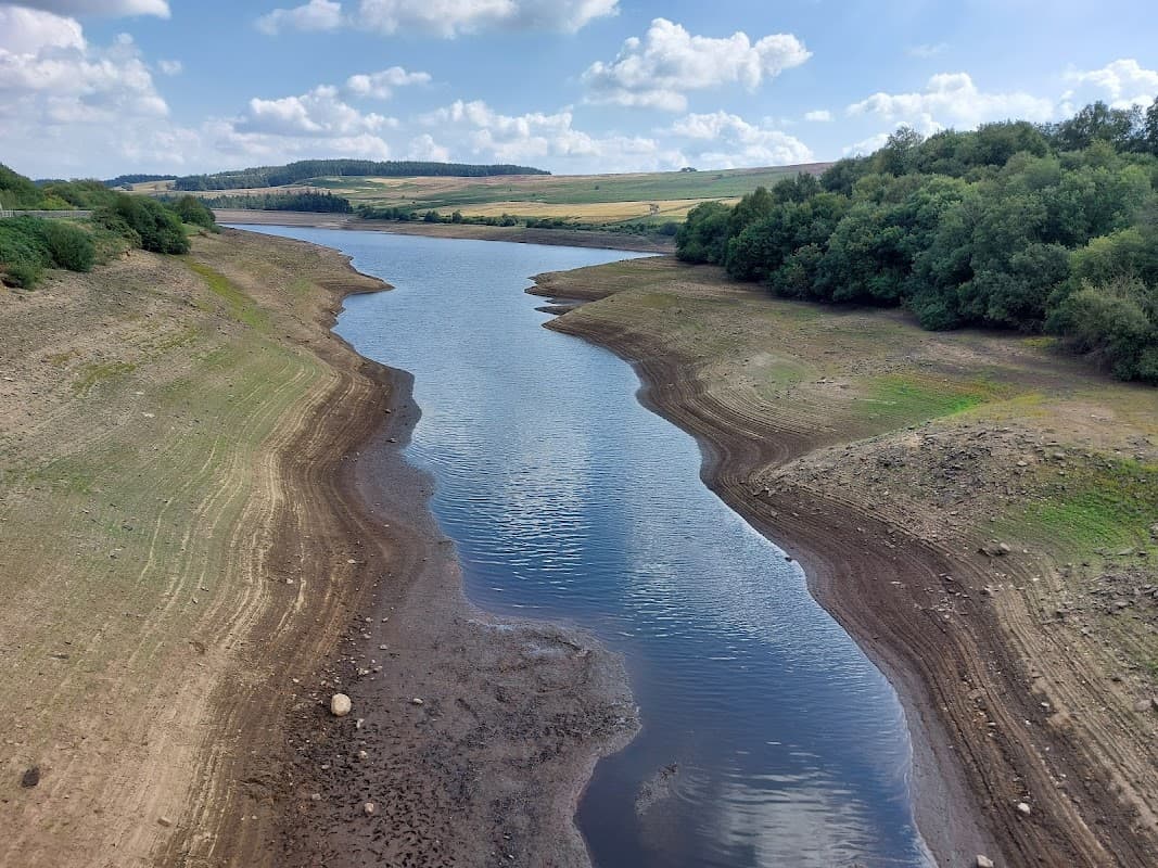 Serene reservoir landscape with calm water, grassy banks, and distant hills under a partly cloudy sky.