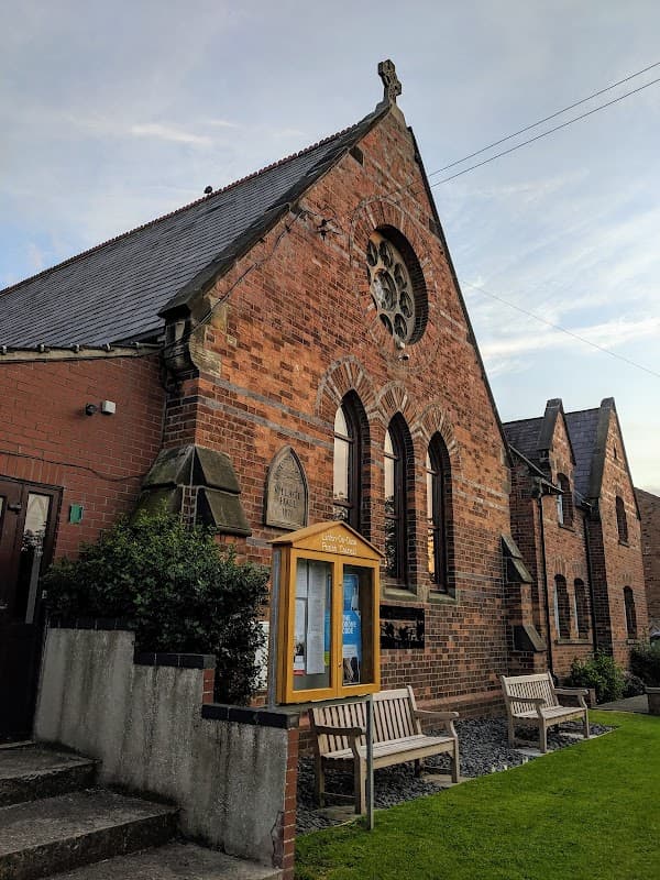 Brick building with arched windows, a decorative rose window, and benches outside in Linton-on-Ouse, Yorkshire.