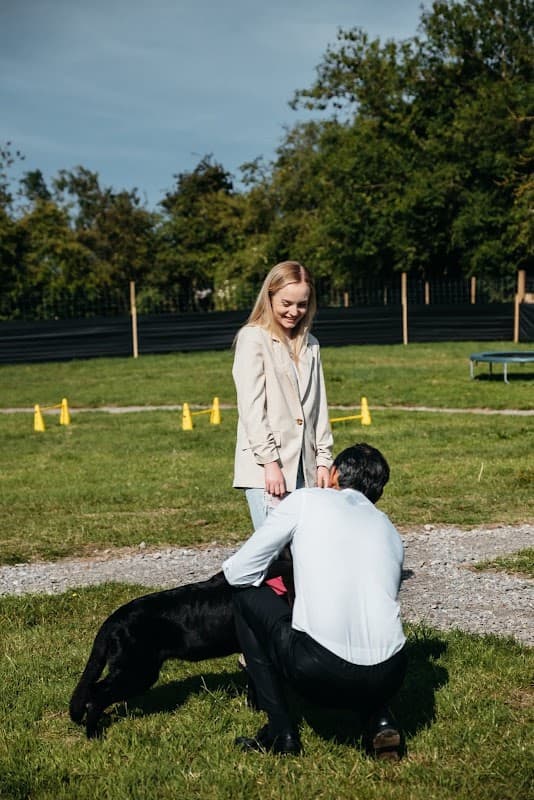 A woman stands with a man kneeling beside a black dog in a grassy area, surrounded by trees and agility cones.