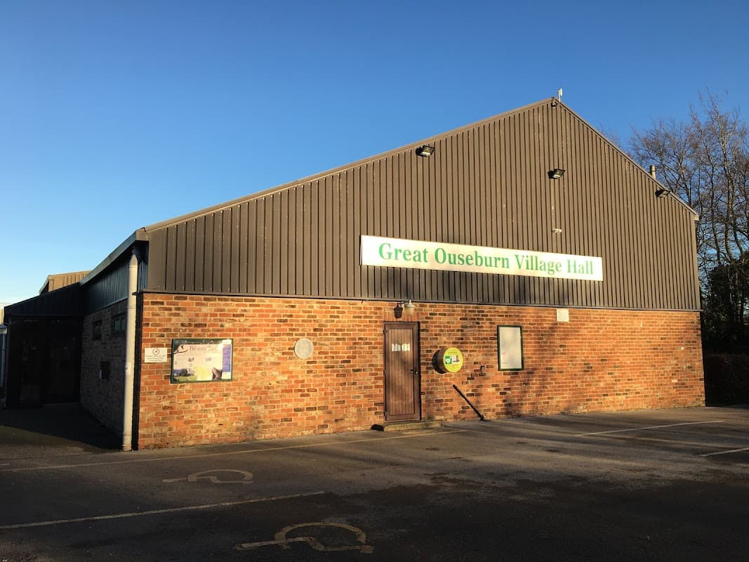 Great Ouseburn Village Hall with a brick facade, metal roof, and signage against a clear blue sky.