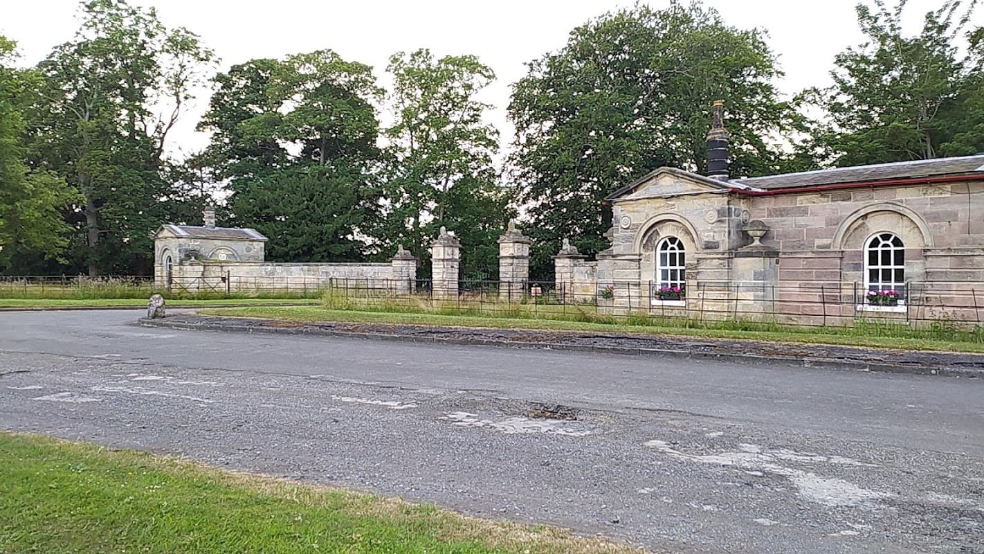 Historic stone buildings with arched windows, surrounded by lush greenery and a gravel path in Little Ribston, Yorkshire.