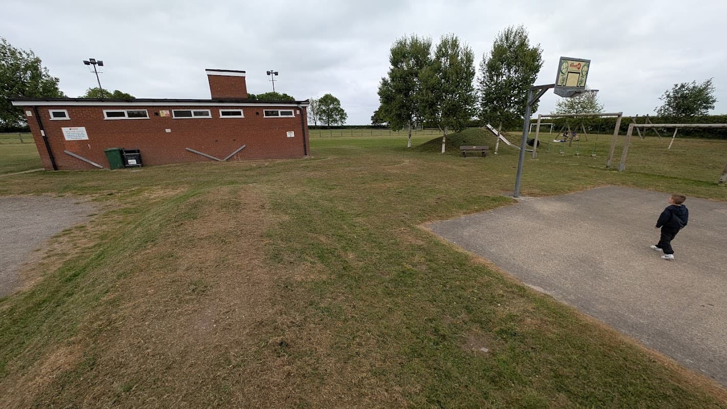 Recreation ground featuring a brick building, basketball hoop, playground equipment, and grassy areas under cloudy skies.