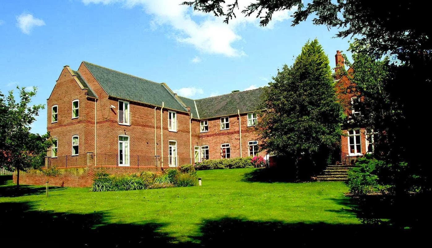 Red-brick manor house surrounded by lush green lawns and trees under a blue sky with scattered clouds.