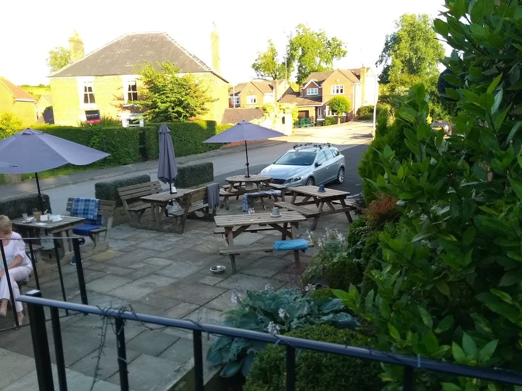 Outdoor seating area with wooden tables and umbrellas, surrounded by greenery, near a quiet street in Little Weighton.