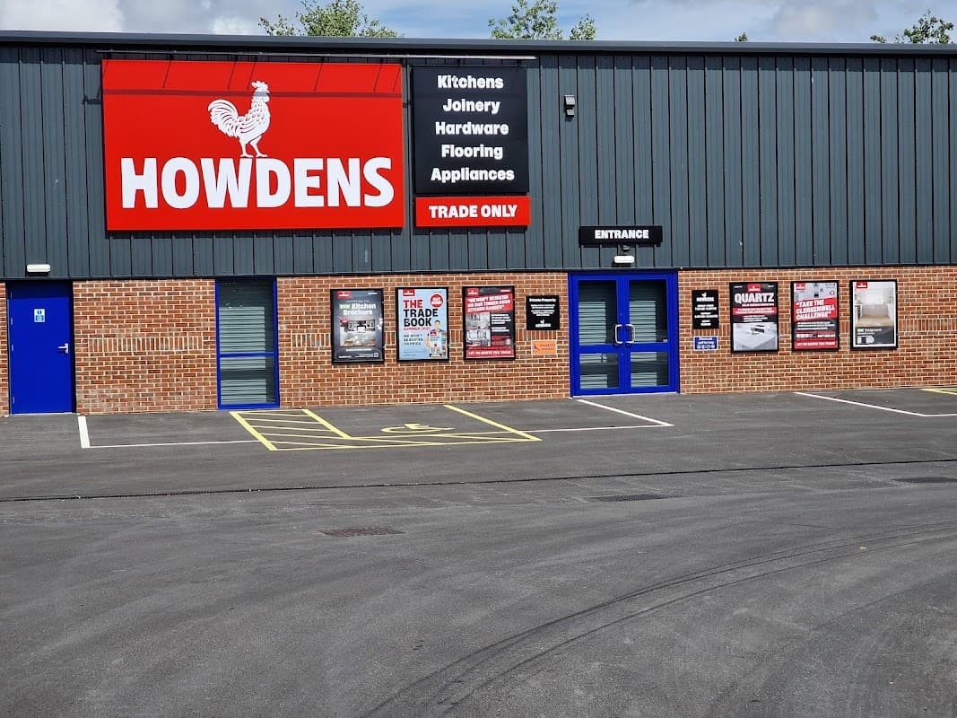Howdens storefront featuring a red sign, blue entrance doors, and promotional posters on a brick wall.
