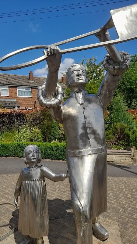 Metal sculptures of a man holding a shovel and a girl, set in a green park with residential buildings in the background.