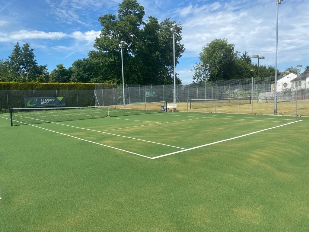 Green tennis court with a net, surrounded by trees and a clear blue sky at Liversedge Tennis Club.